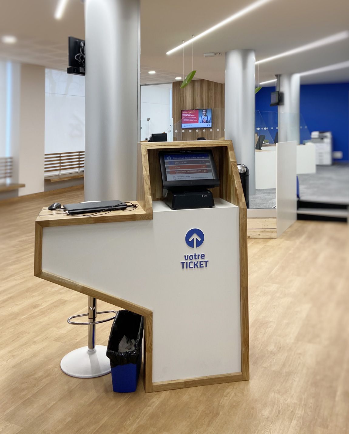 White ticket kiosk with a screen, logo, and a bar stool inside a building.