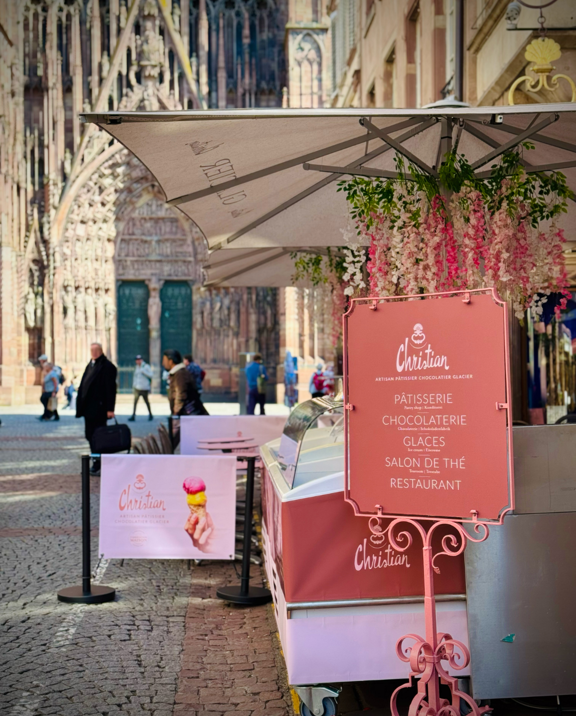 Stand de glaces rose avec enseigne, fleurs et cathédrale strasbourg en arrière-plan.