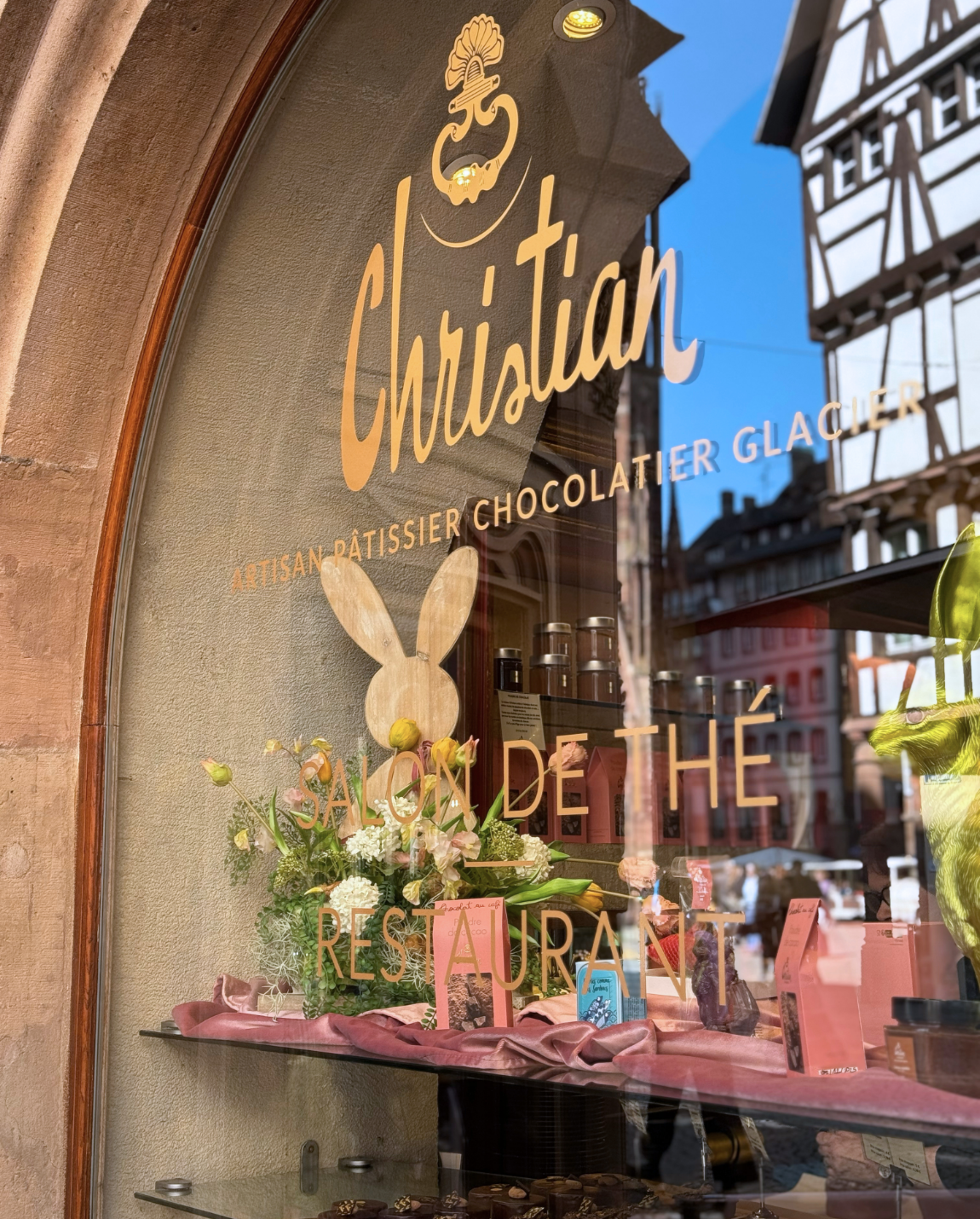 Vitrine de magasin avec inscription dorée « Christian », décor de lapin de Pâques et maisons à colombages.