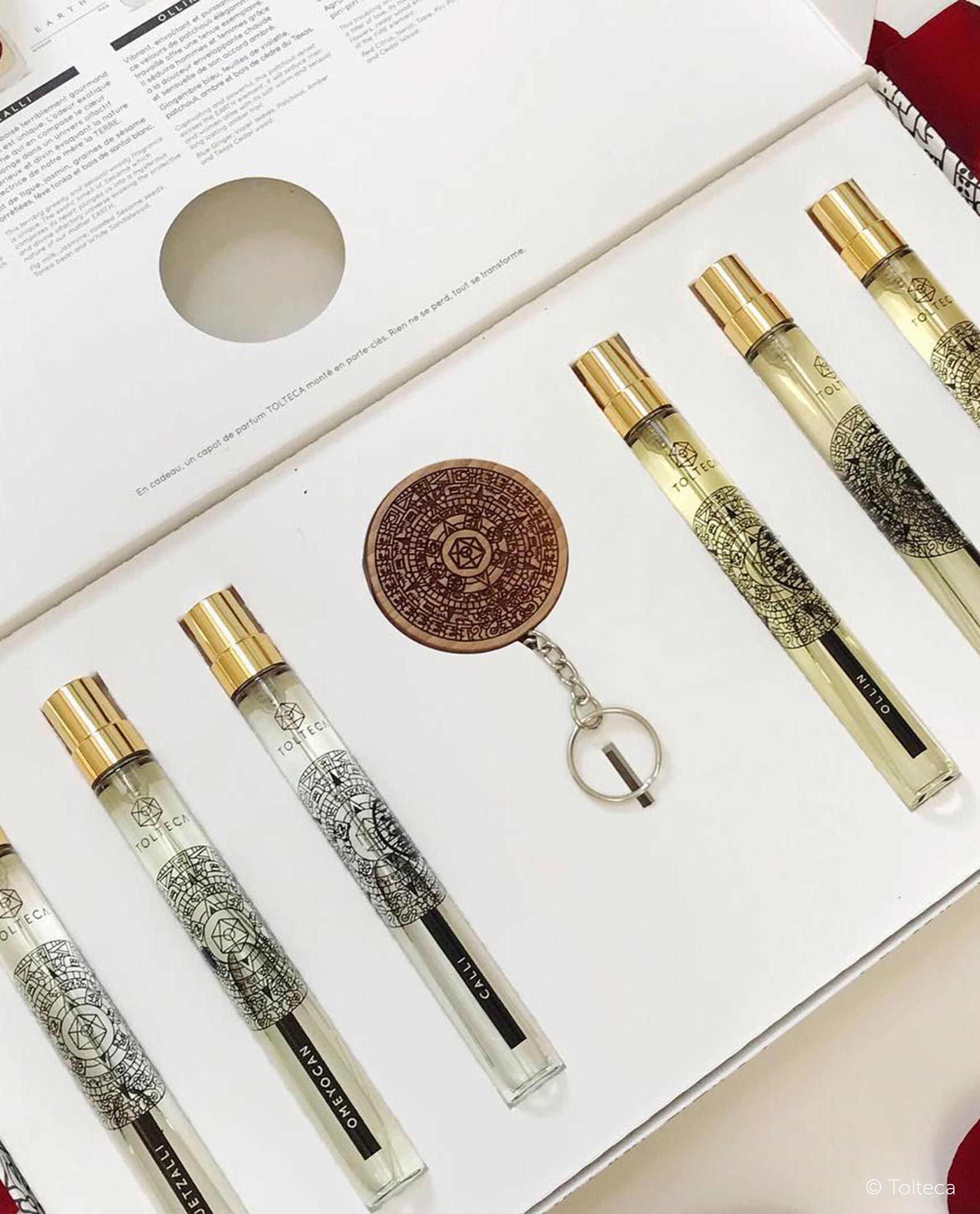 Open box with six perfume vials and a round keychain. Gold accents, white background.