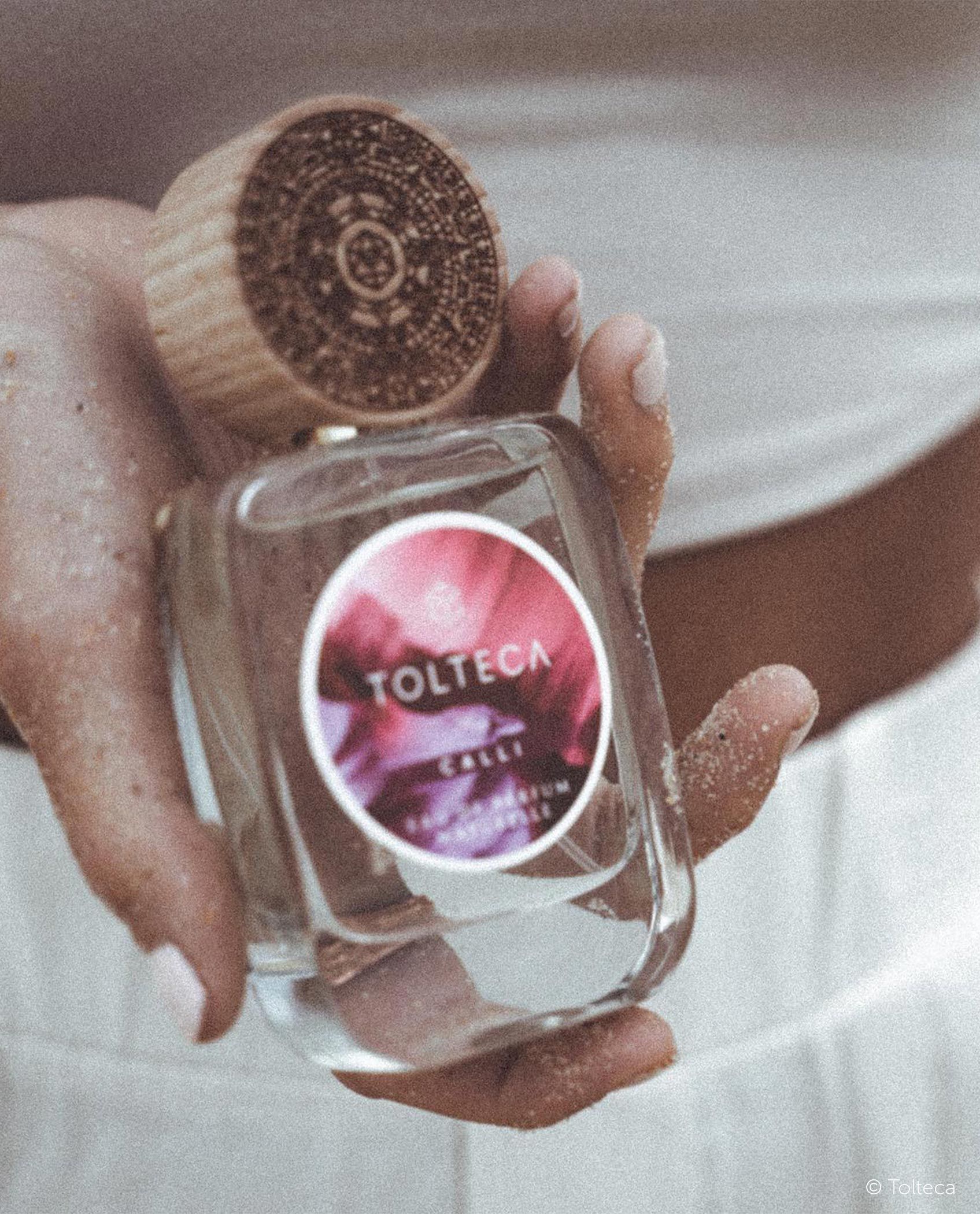 A clear glass perfume bottle with a wooden cap, held by a person in a soft, white setting.