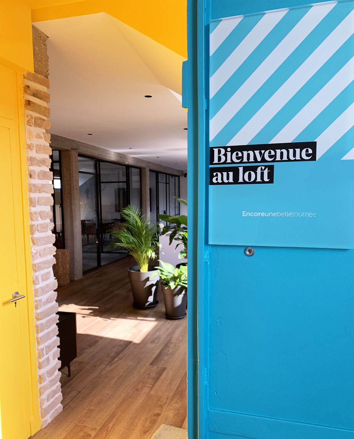 Porte bleue ouverte avec l'inscription « Bienvenue au loft ». Un mur jaune et des briques encadrent le couloir intérieur.