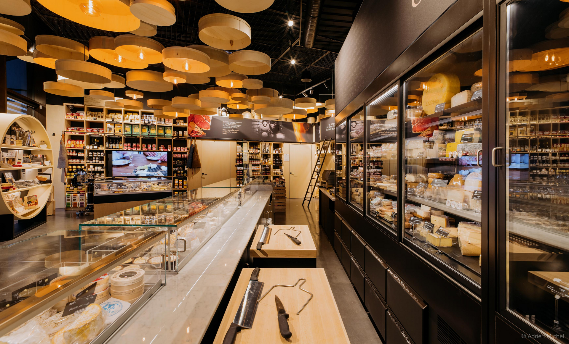 Interior of a cheese shop with display cases, a long counter, and overhead wooden discs for lighting.