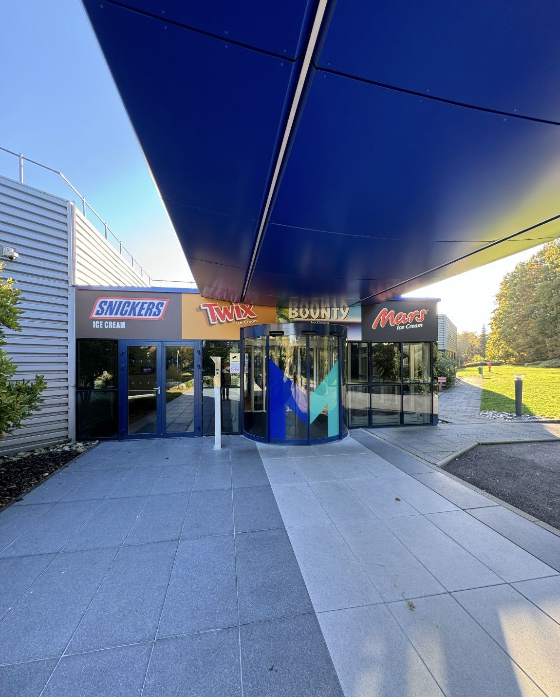 Entrance to a building under a blue canopy. Signage features several businesses. Paved walkway leads to glass doors.