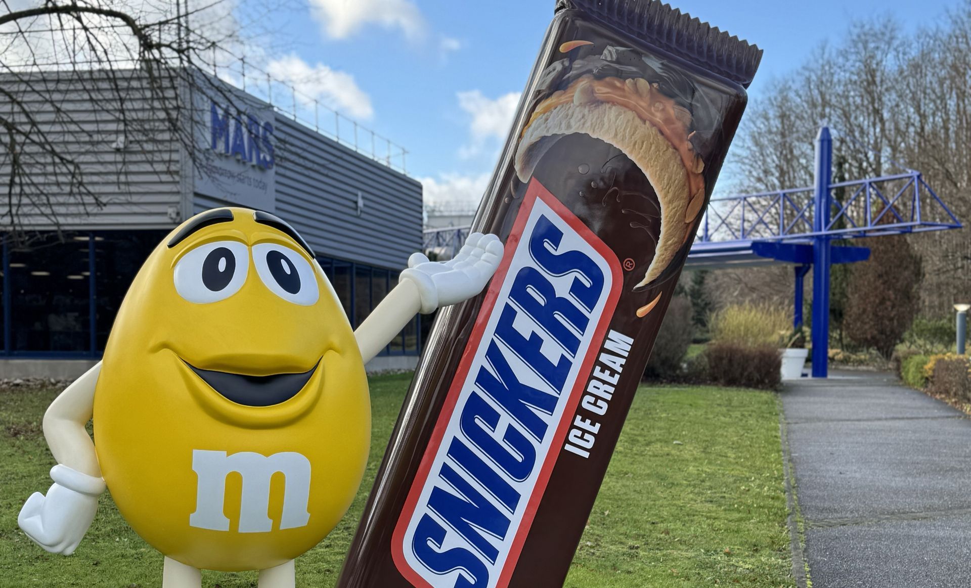 Yellow M&M character with Snickers ice cream bar outside Mars factory.