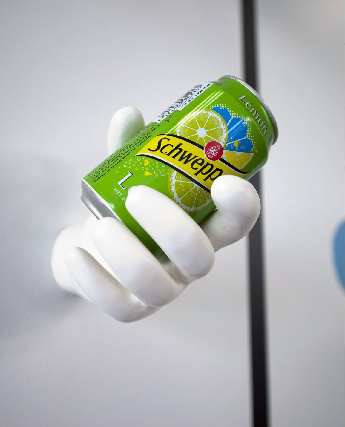 A white hand sculpture holding a green Schweppes can against a white wall.