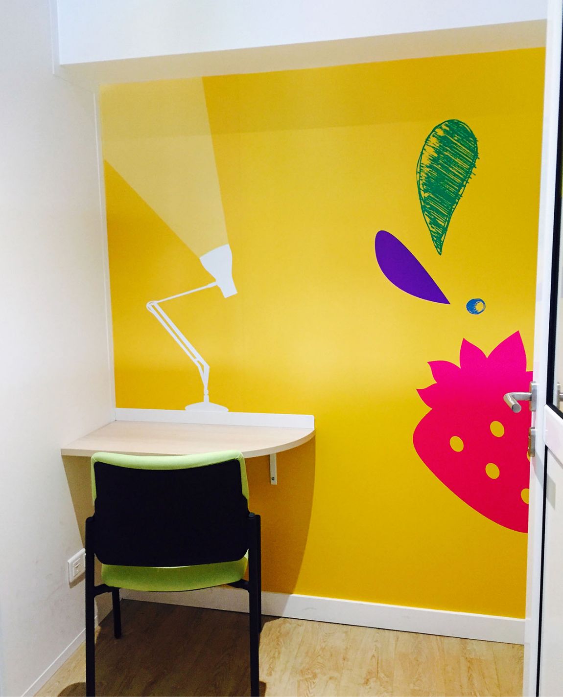 A brightly colored workspace. Yellow wall with desk, chair, and cartoon art of lamp and fruit. White door frame.