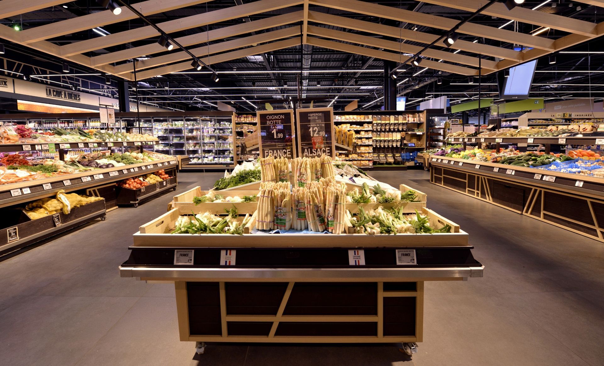 Allée d'un supermarché avec un présentoir central de fruits et légumes, un éclairage puissant et des étagères remplies de fruits et légumes.