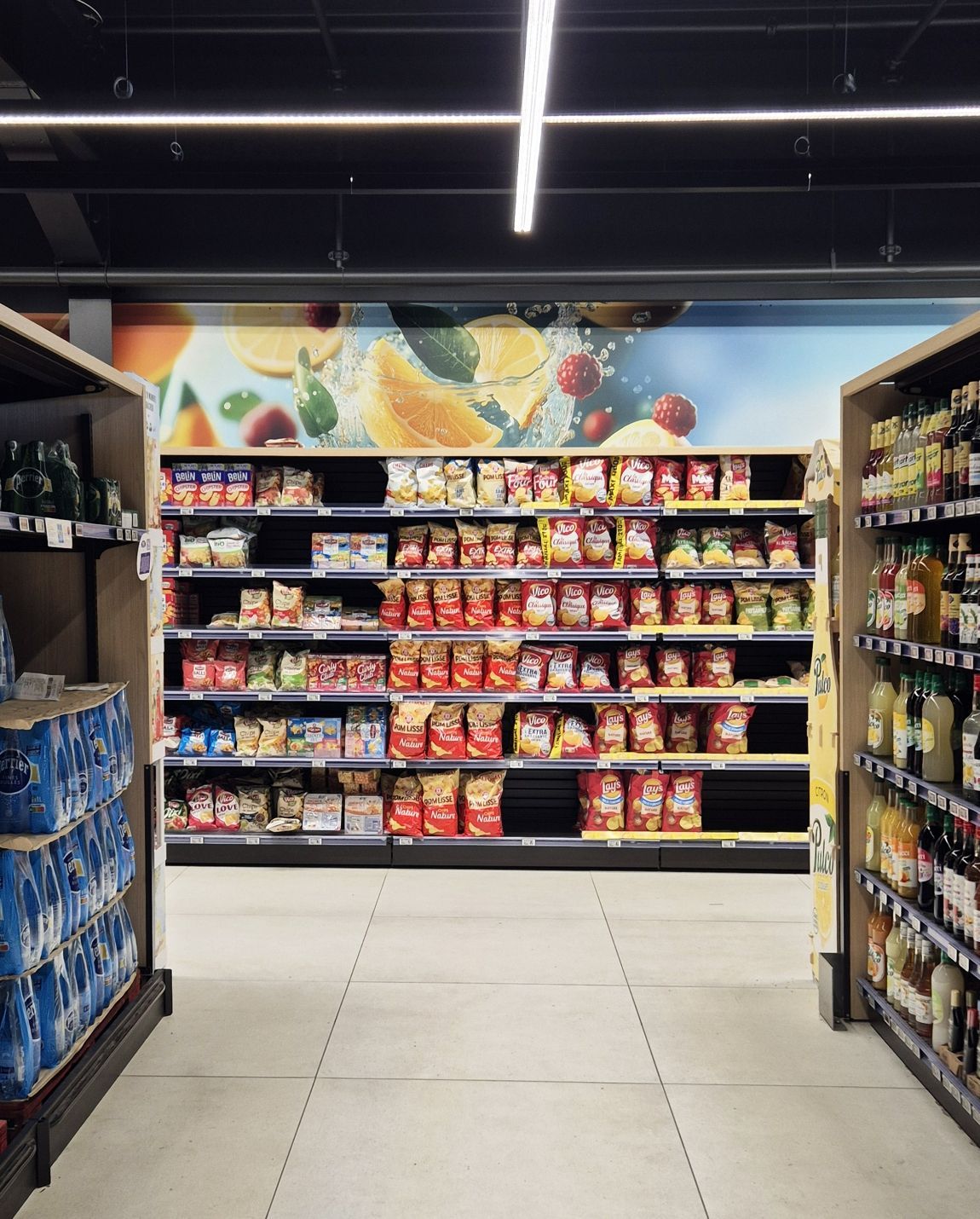 Allée d'un supermarché avec des étagères de snacks et de boissons éclairées par des néons.