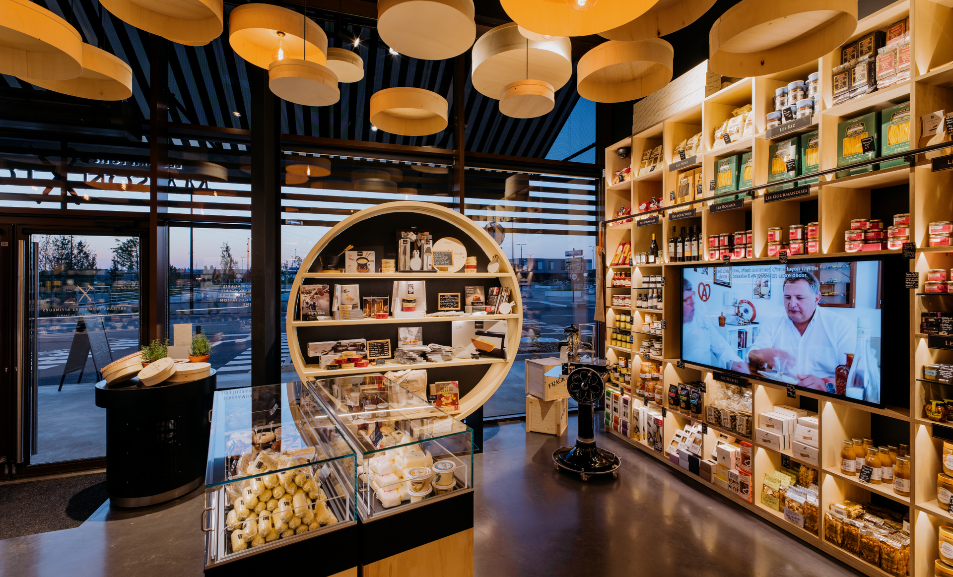 Interior shot of a specialty food store with shelves of products, a display case, and a screen showing a chef.
