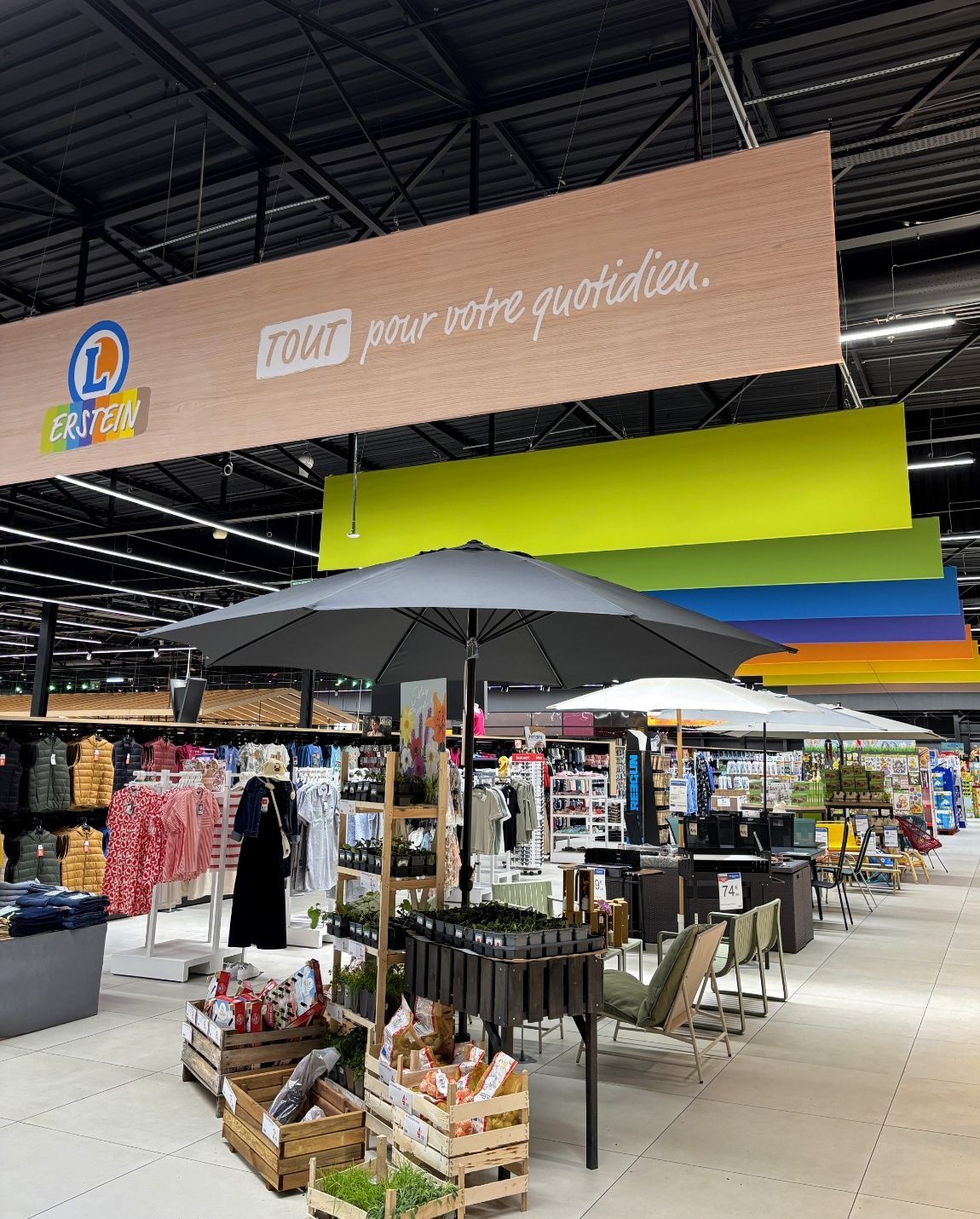 Marché couvert avec parasols, portants à vêtements et produits frais sous des enseignes suspendues colorées