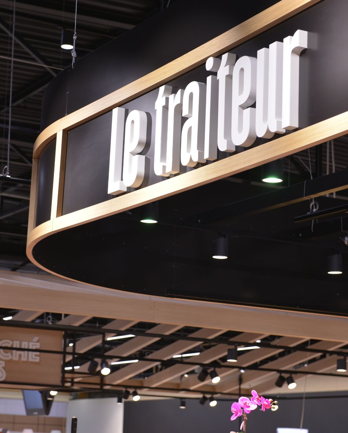 Enseigne noire suspendue au-dessus d'un marché couvert moderne ou d'une halle alimentaire, indiquant « Le Traiteur ».