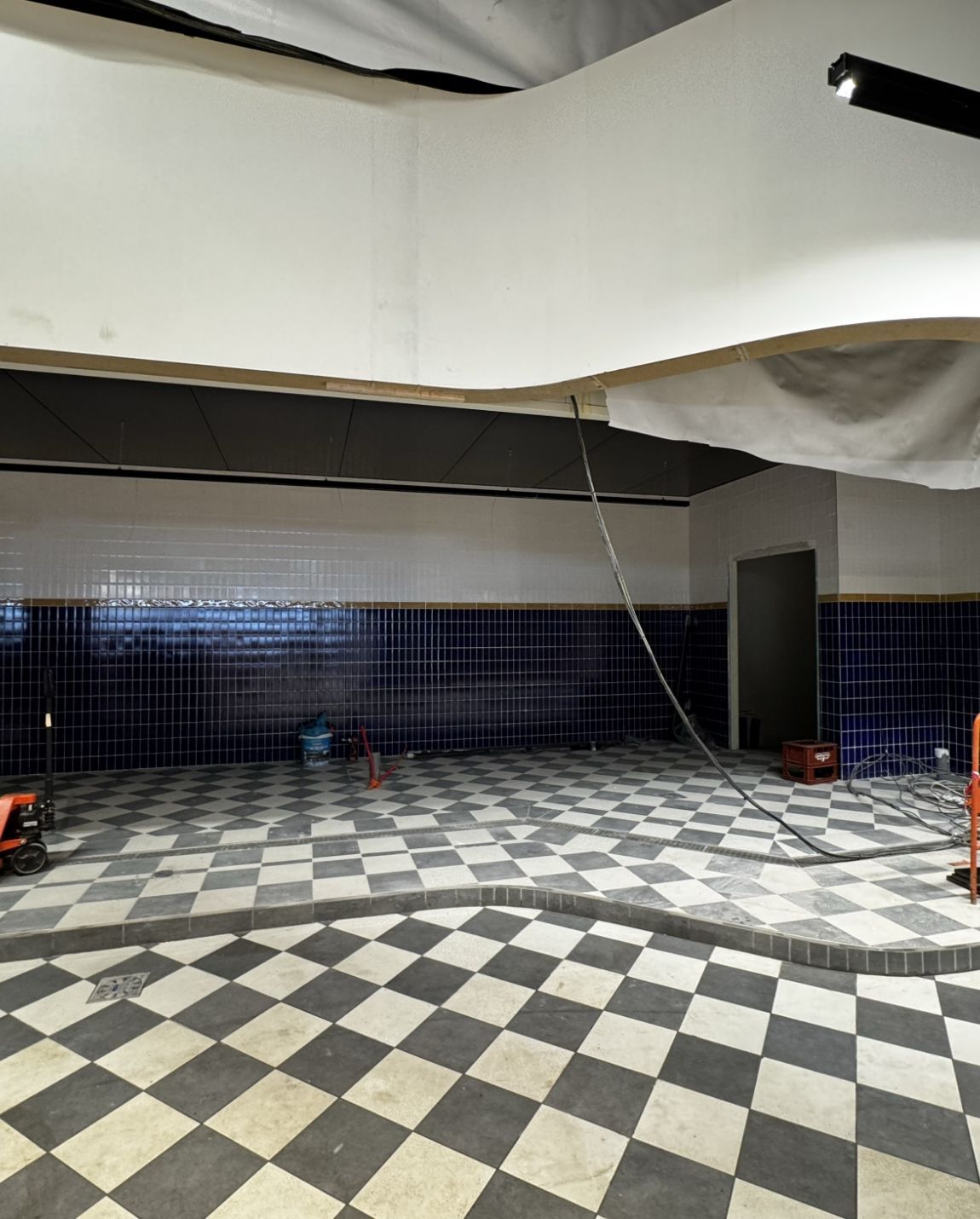 Travaux hypermarché, sol à damier noir et blanc, murs carrelés sombres et un câble suspendu au-dessus.