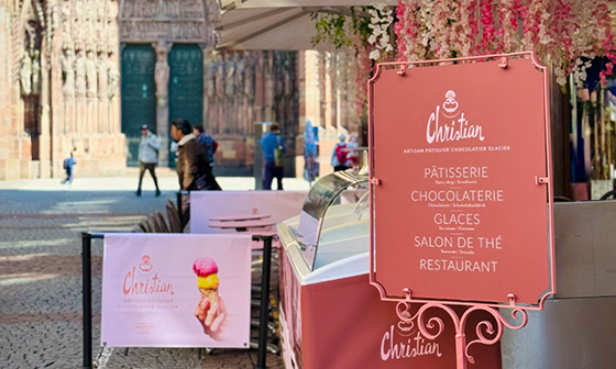 Pink and white Christian pastry shop stand in a plaza with people and cathedral in the background.