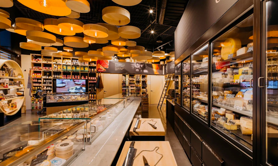 Intérieur d'une épicerie fine avec présentoir à fromages, plafonniers en bois et divers produits alimentaires.