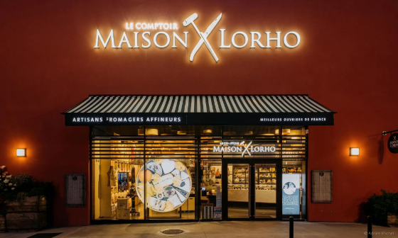 Boutique Maison Lorho avec enseigne lumineuse ; façade rouge, toit noir. Vitrines vitrées pour la présentation des produits.