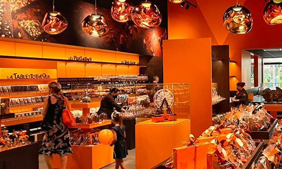 Intérieur d'une chocolaterie lumineuse avec des clients. Présentoirs, décoration orange et luminaires en cuivre.
