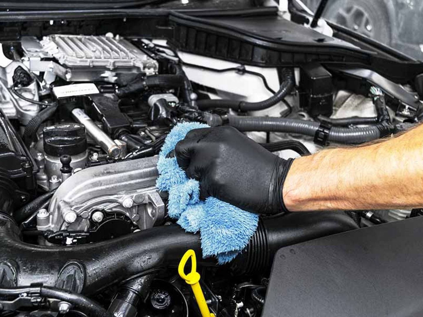 Gloved hands wiping car engine components with a blue cloth in the engine bay