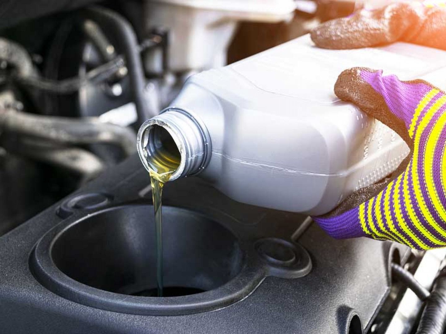 Gloved hand pouring motor oil from a white bottle into an engine fill port