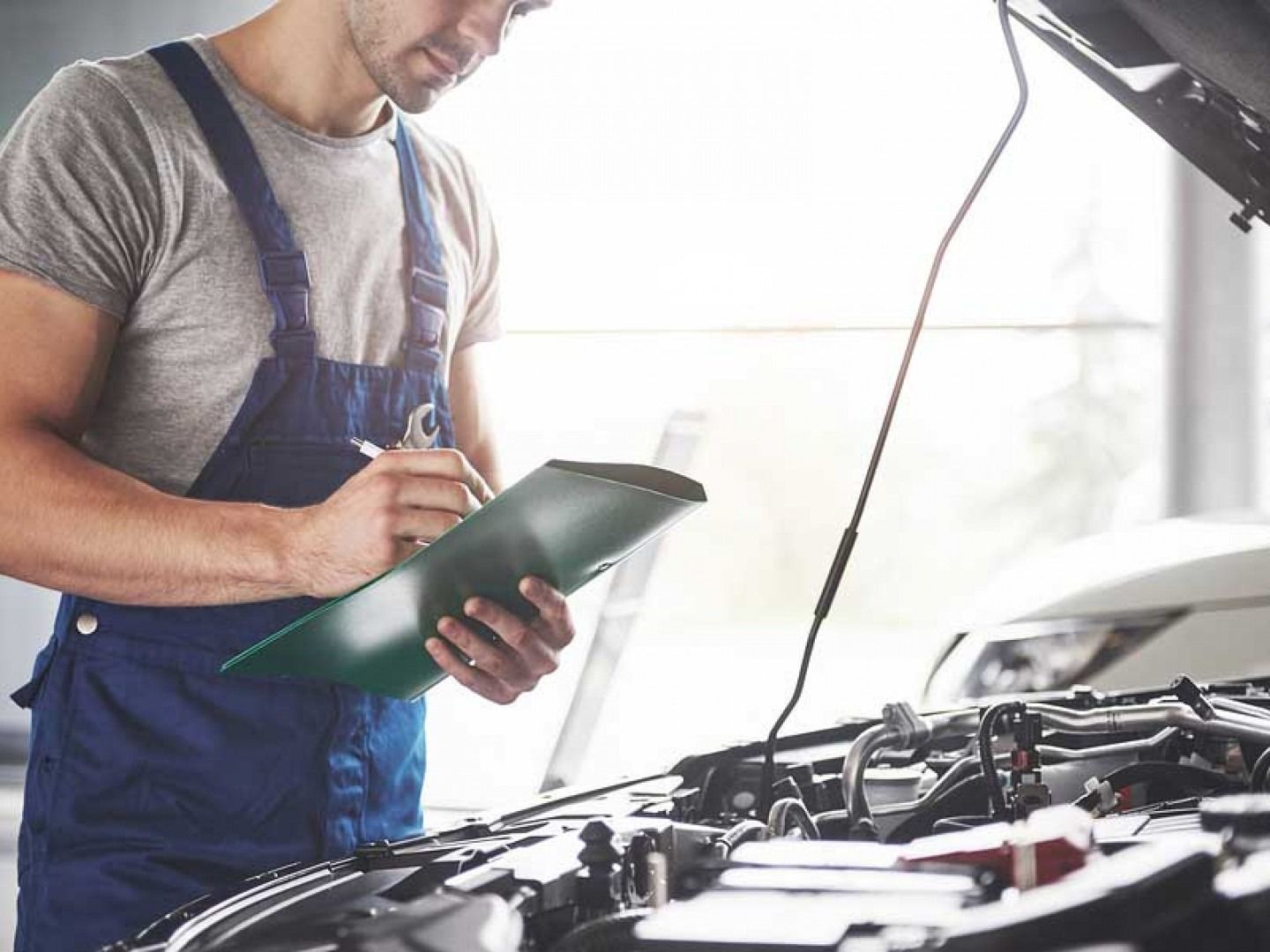 Mechanic in blue overalls inspecting a car engine with a clipboard in a garage