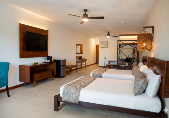 Habitación de hotel luminosa y moderna con dos camas individuales, televisor, escritorio, ventilador de techo y lámpara de mimbre.