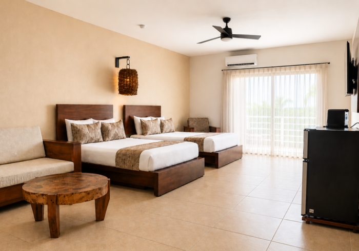 Amplia habitación beige con dos camas individuales, una mesa redonda de madera, ventilador de techo y puertas corredizas de cristal.