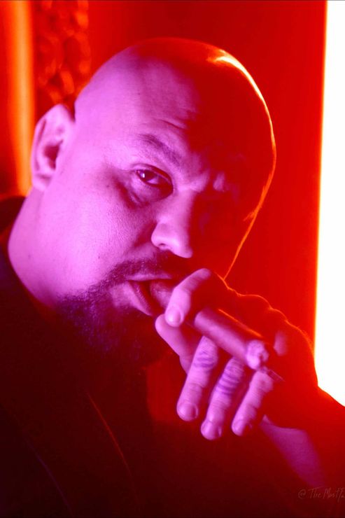 Bald man in red lighting, resting his hand near his face, looking at the camera.