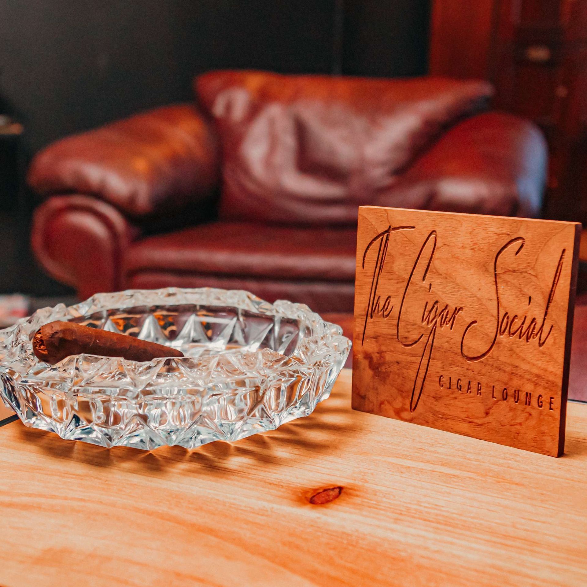 Crystal ashtray with cigar on a wooden table beside a signed wooden sign, with a red leather chair in the background