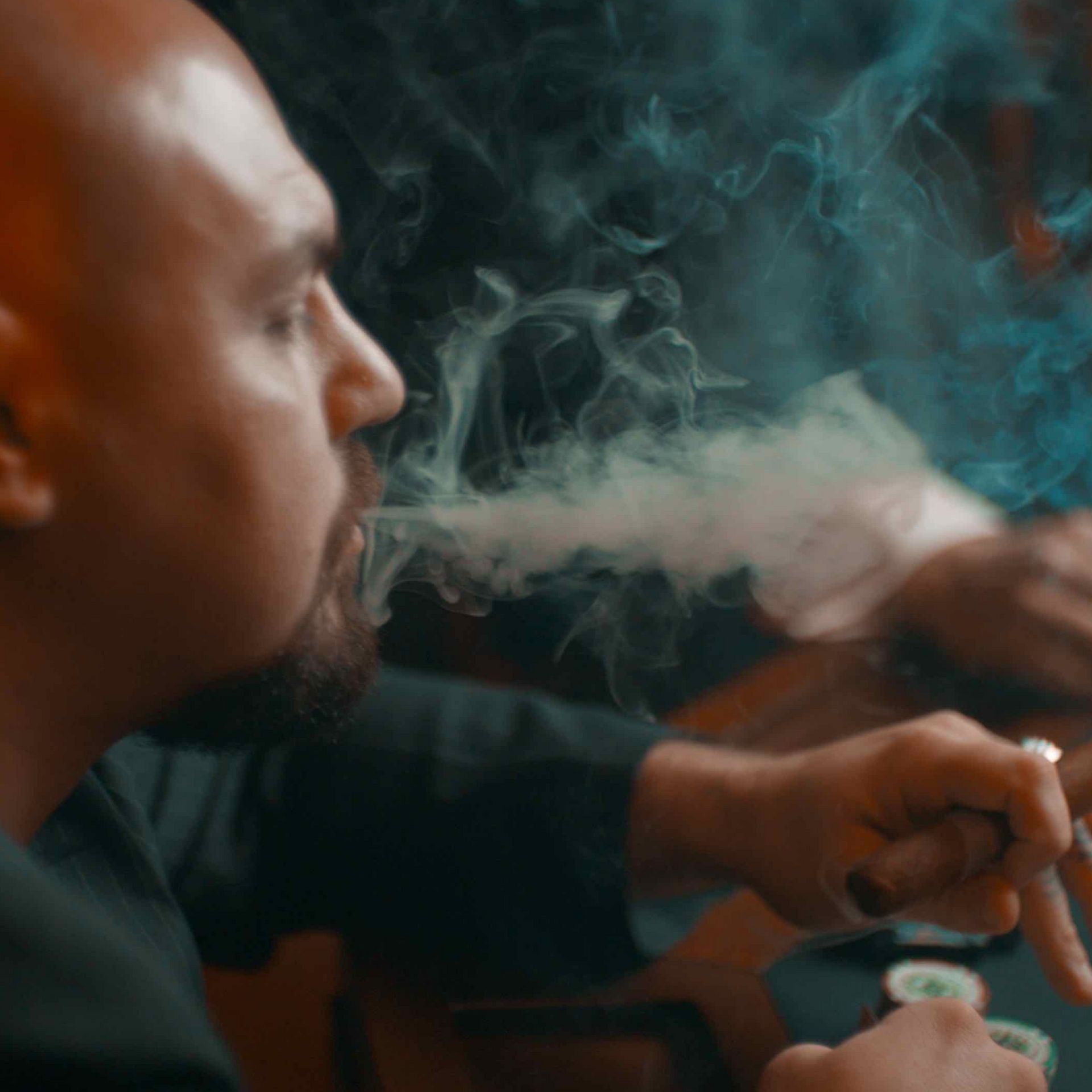 Man exhaling smoke indoors, close-up side profile with a blurred hand holding a cigarette.
