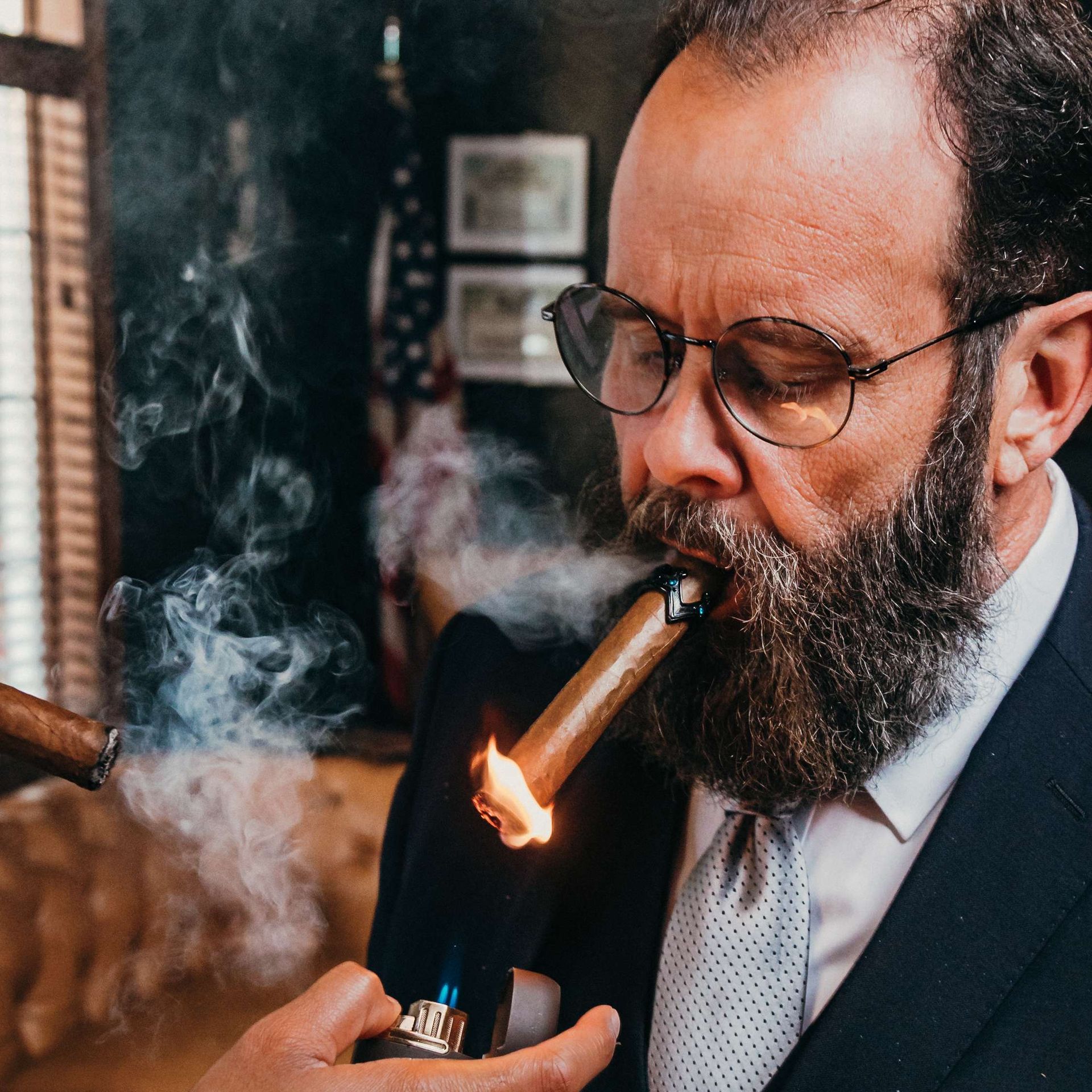 Man in glasses smoking a lit cigar, with smoke swirling around him indoors.
