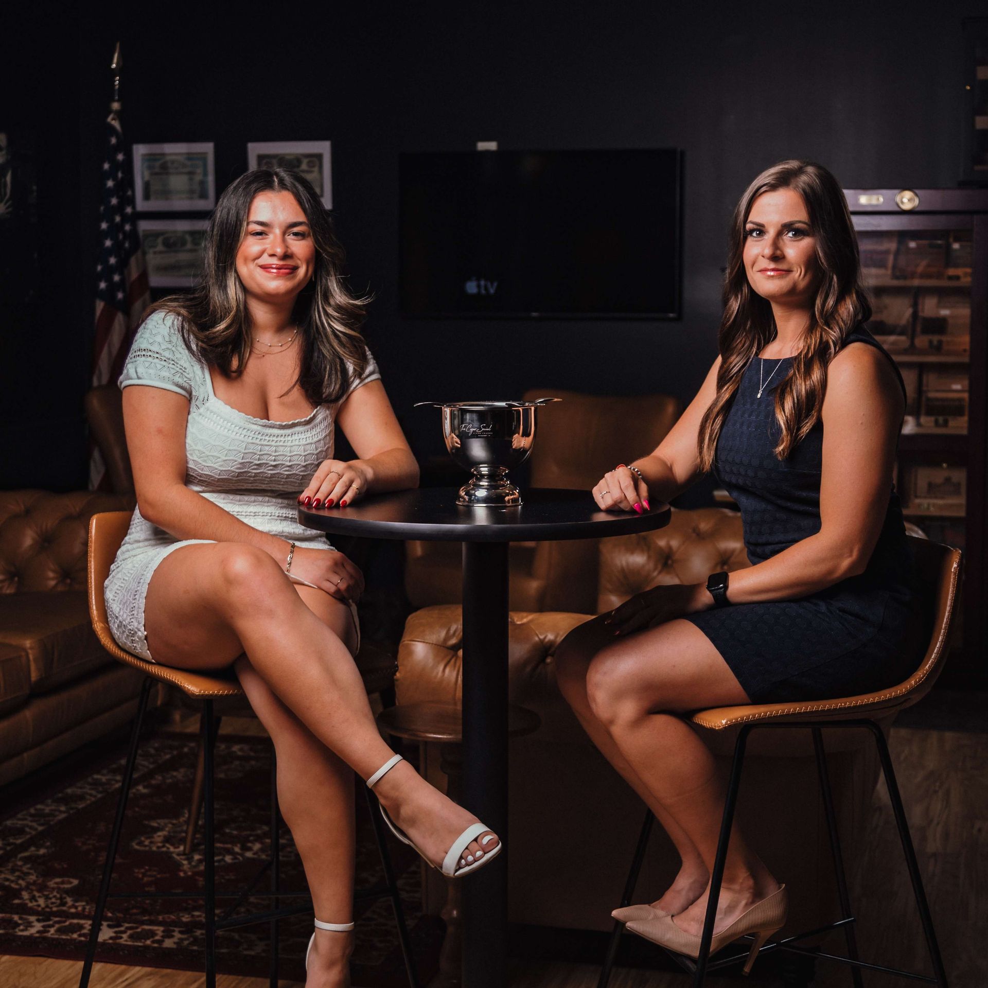 Two women sit at a small table in a dimly lit bar, smiling toward the camera.