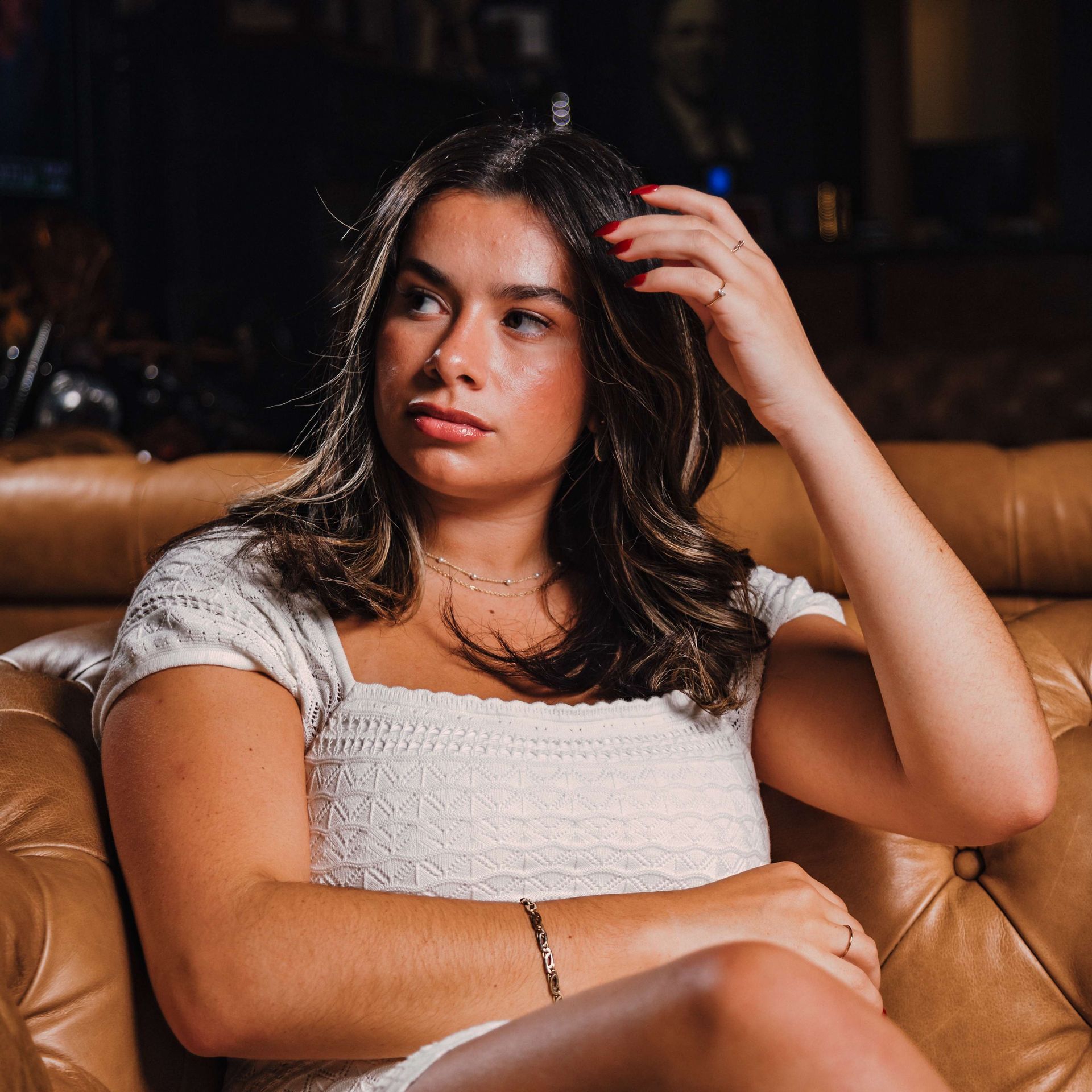 Woman with dark hair seated on a tan couch, looking aside with one hand in her hair.