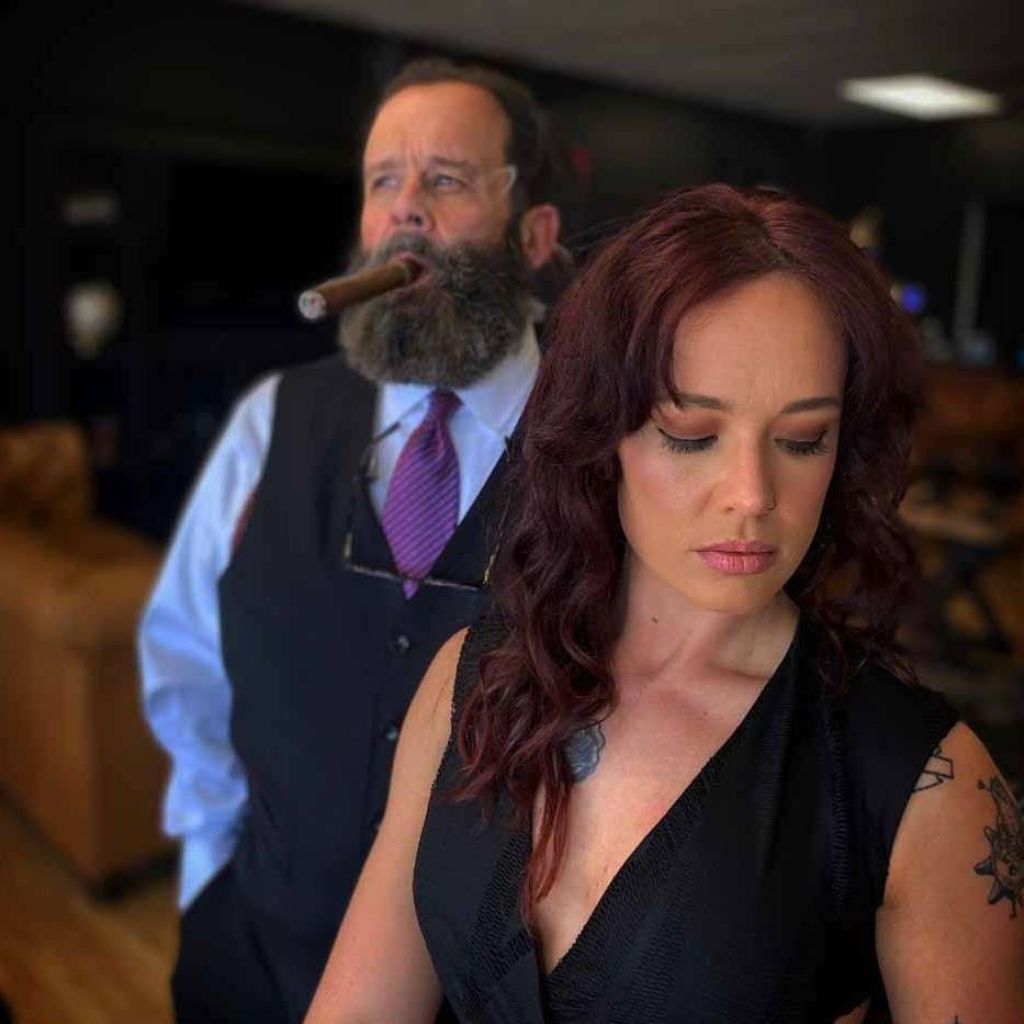 Man in vest smoking a cigar behind a woman with red hair in a dark dress, indoors with a blurred background