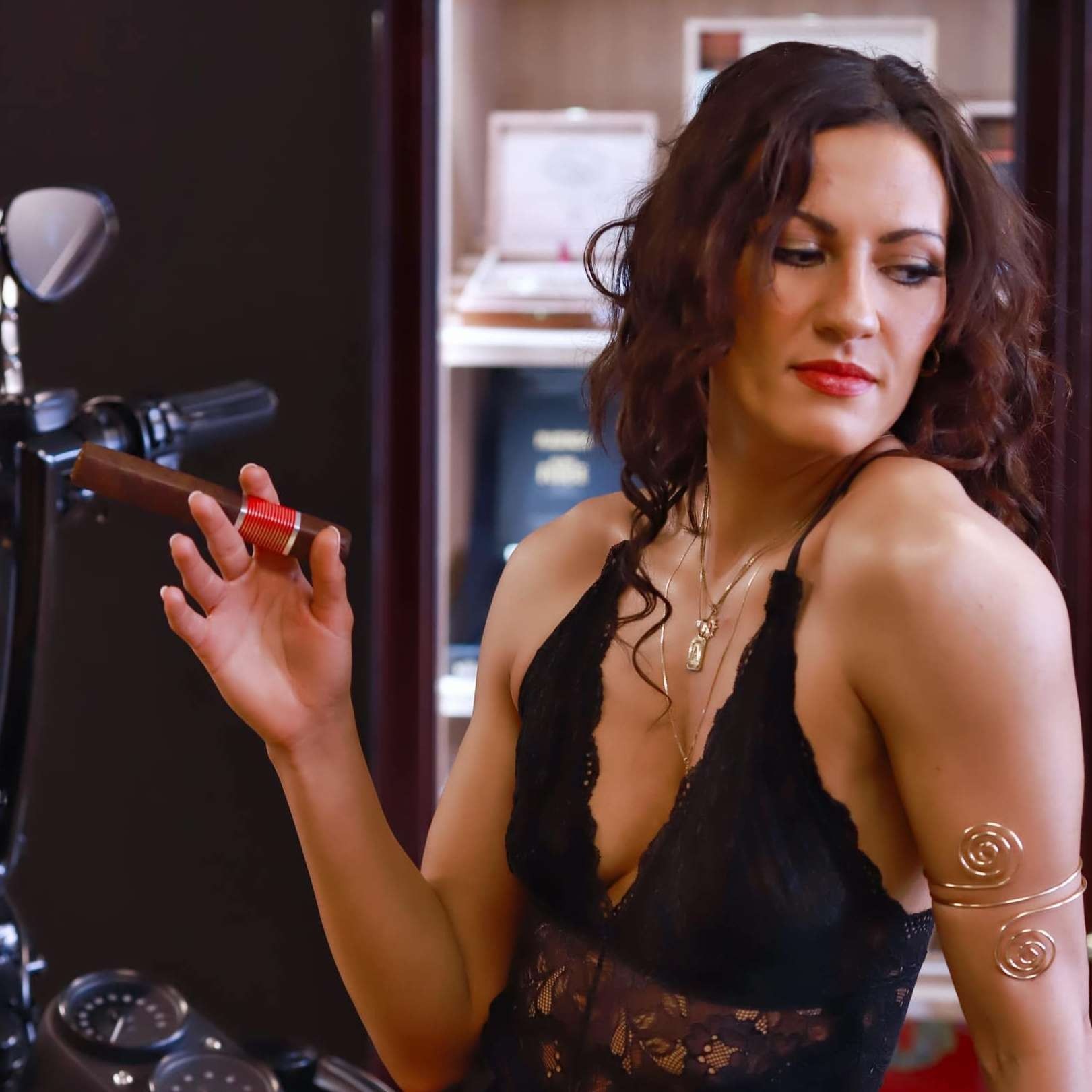 Woman in a black lace top holding lipstick beside a vanity, looking over her shoulder.