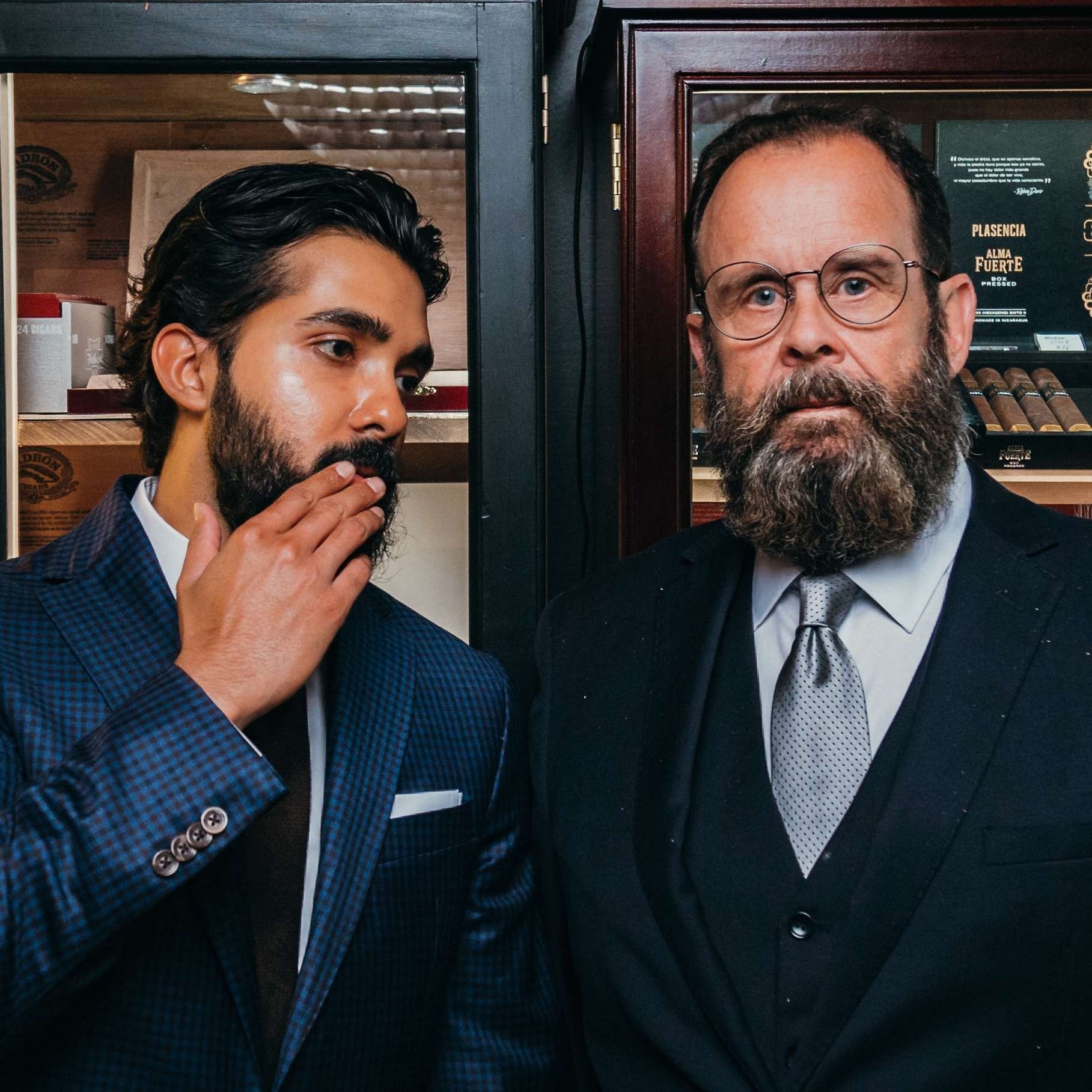 Two men in suits stand indoors, one speaking quietly to the other near a doorway.
