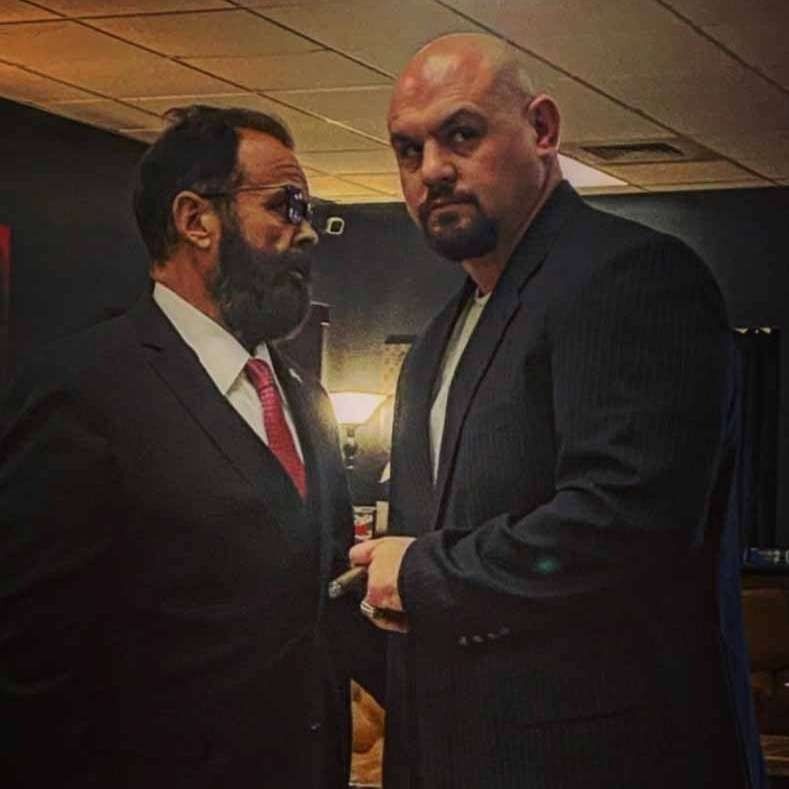 Two suited men stand face-to-face in a dimly lit room, looking serious.