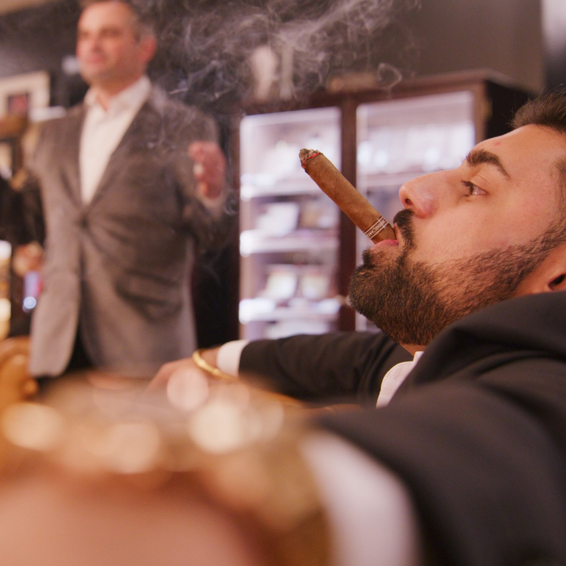Man lounging in a cigar lounge, smoking a cigar while a blurred man stands in the background.