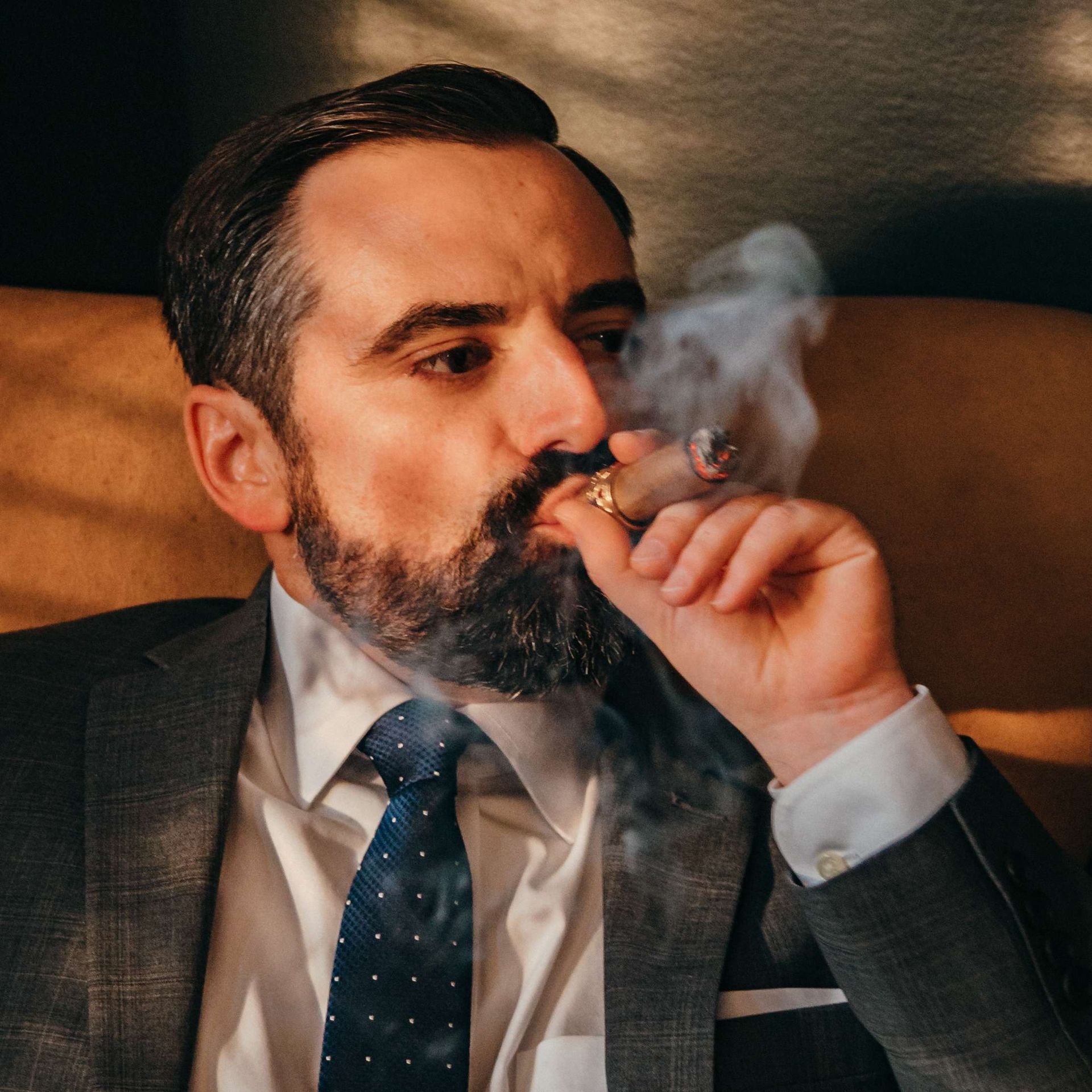 Man in a suit smoking a cigar, exhaling smoke in a dimly lit setting