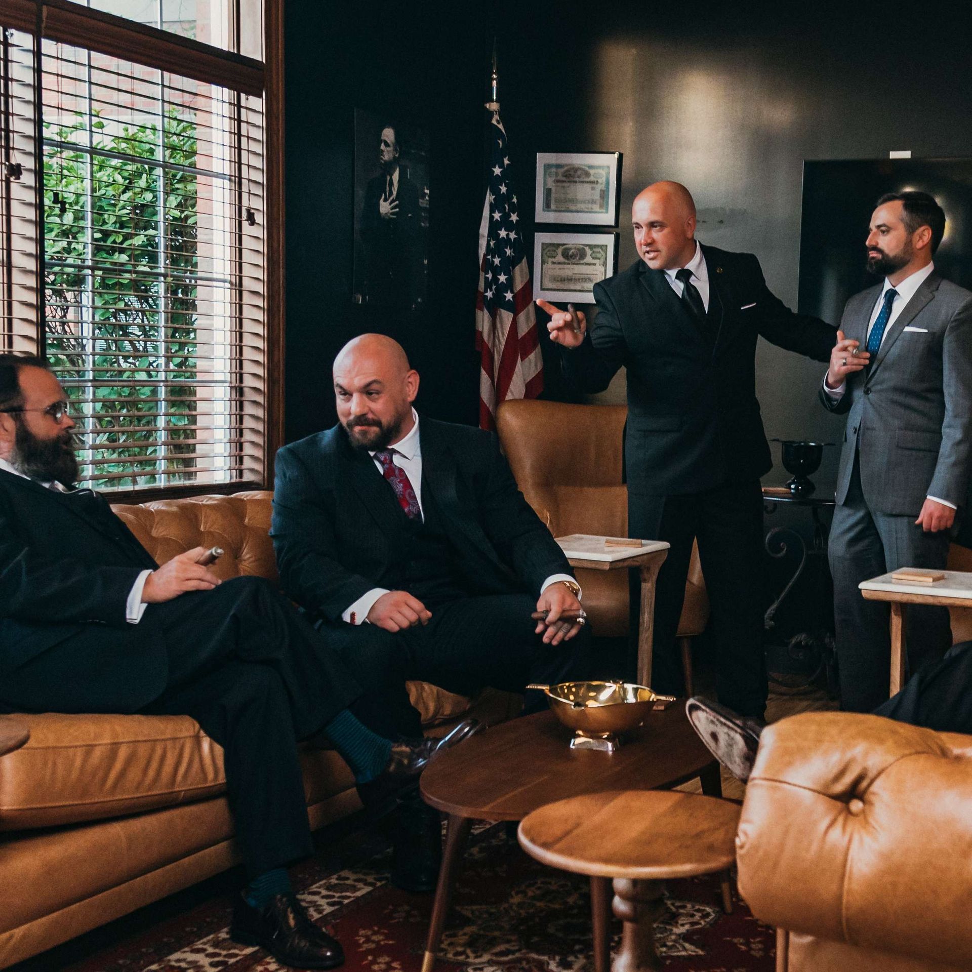Four men in suits talk in a dim office lounge with leather chairs and an American flag.