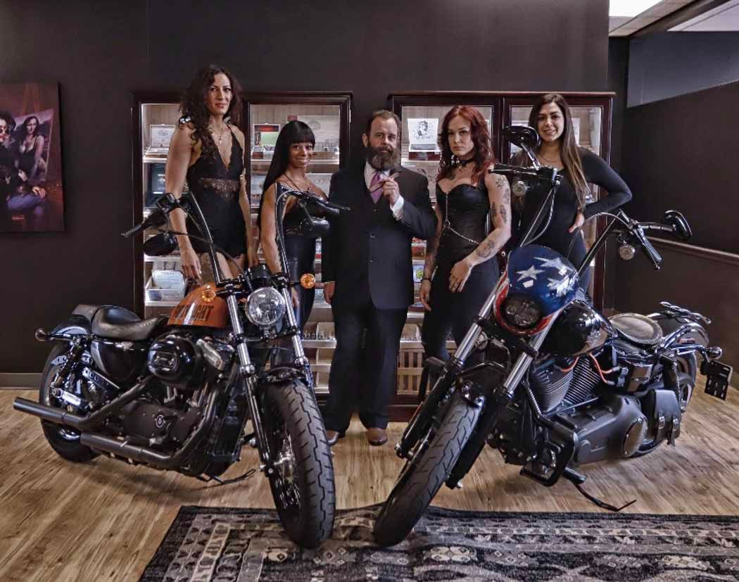 Five people posing with two motorcycles in a showroom, with a dark wall and wooden floor.