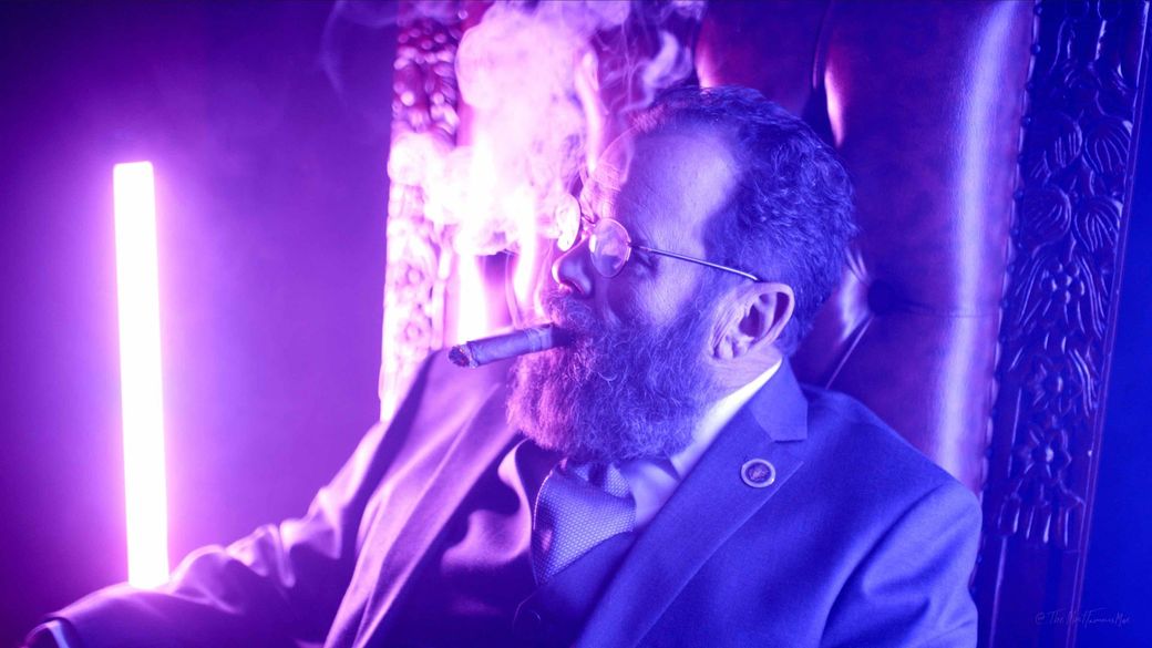 Man in a suit smoking a cigar under purple neon lighting, seated in profile.