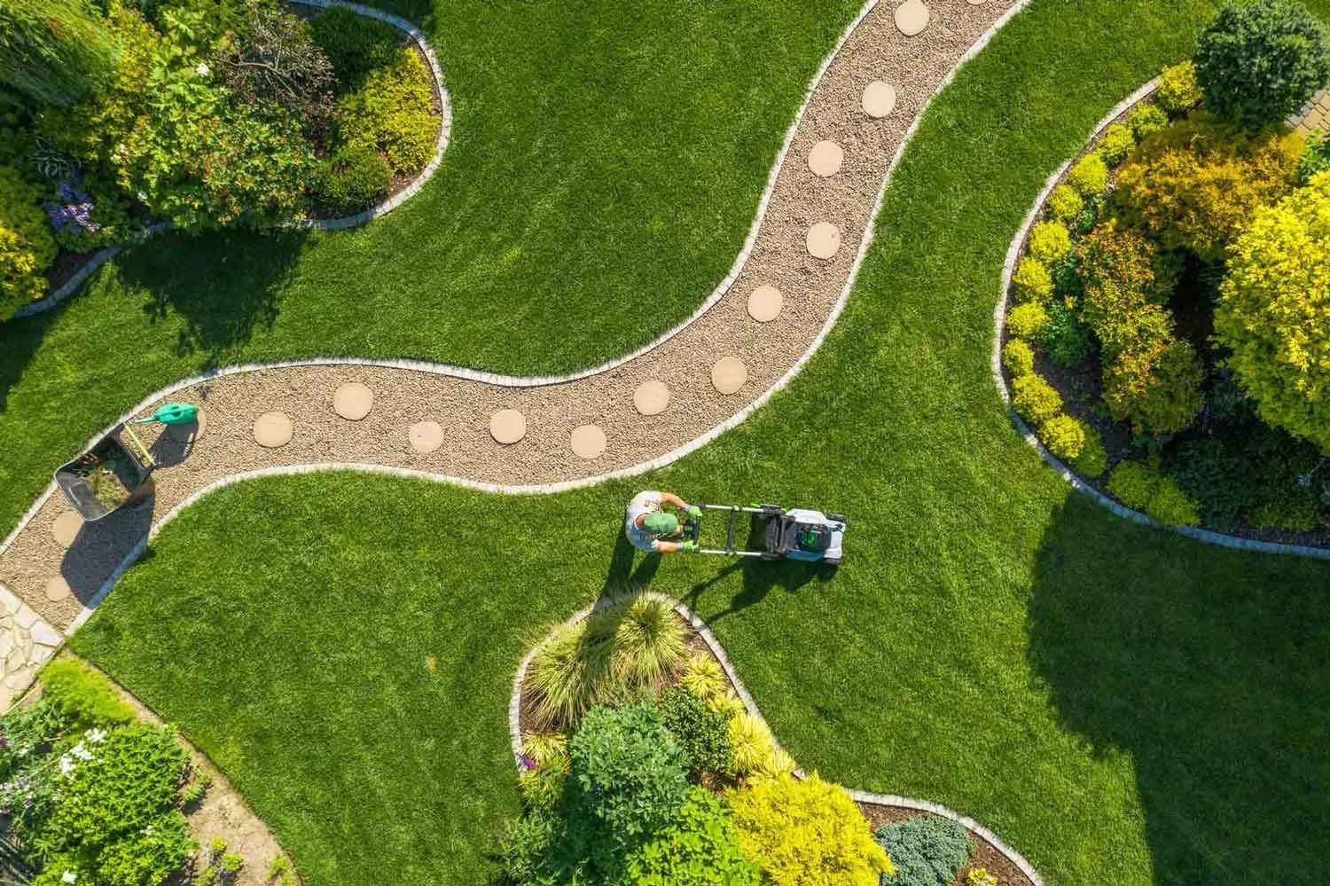 Overhead view of a person mowing a lush green lawn with a curved stone pathway through it, surrounded by colorful gardens.