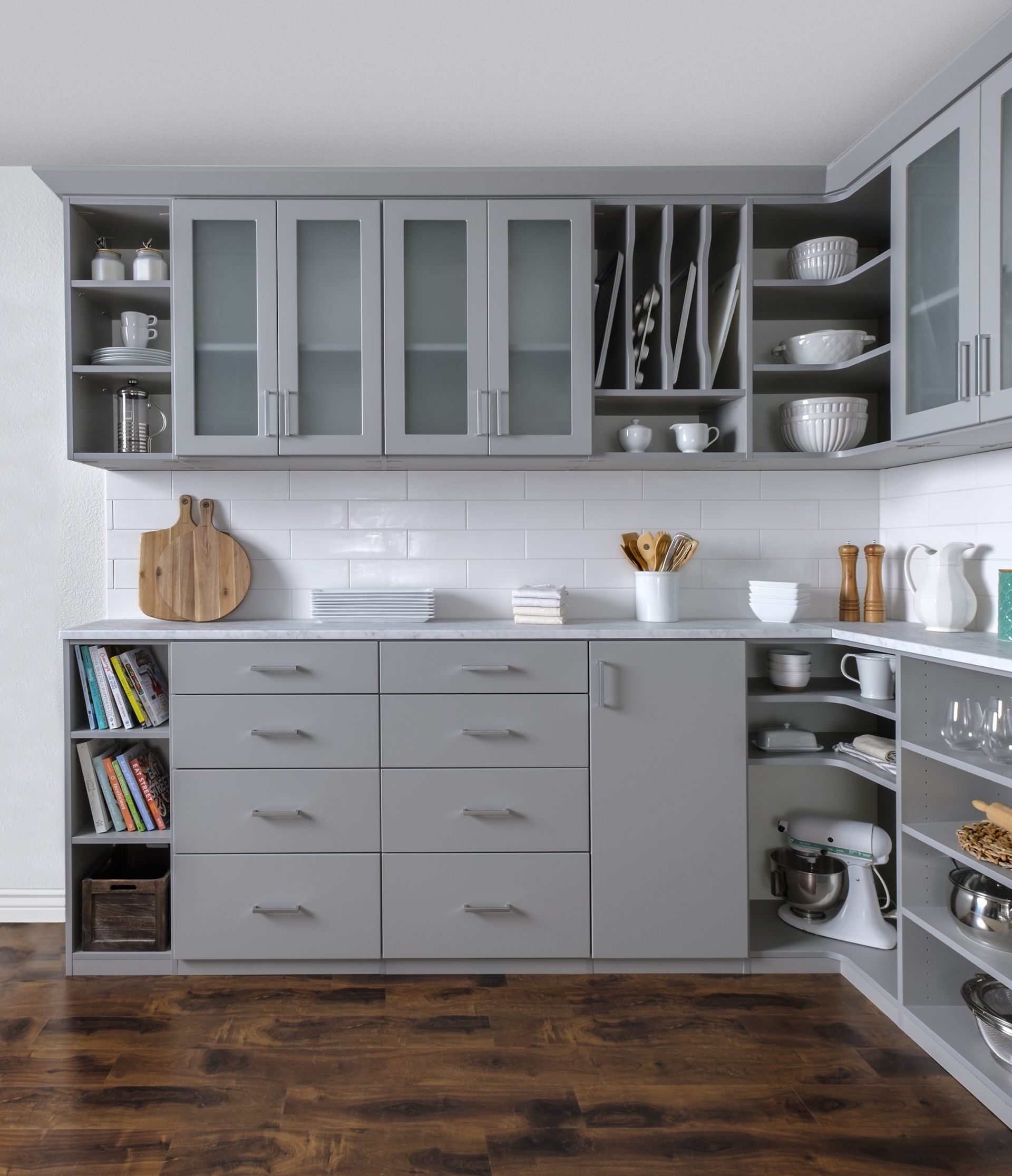 Gray kitchen cabinets with white subway tile backsplash, featuring open shelving, glass-front doors, and a stand mixer.