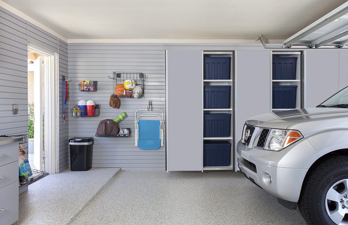 A garage with gray slat walls, storage shelves, blue bins, a black trash can, and a silver car parked on a speckled floor.