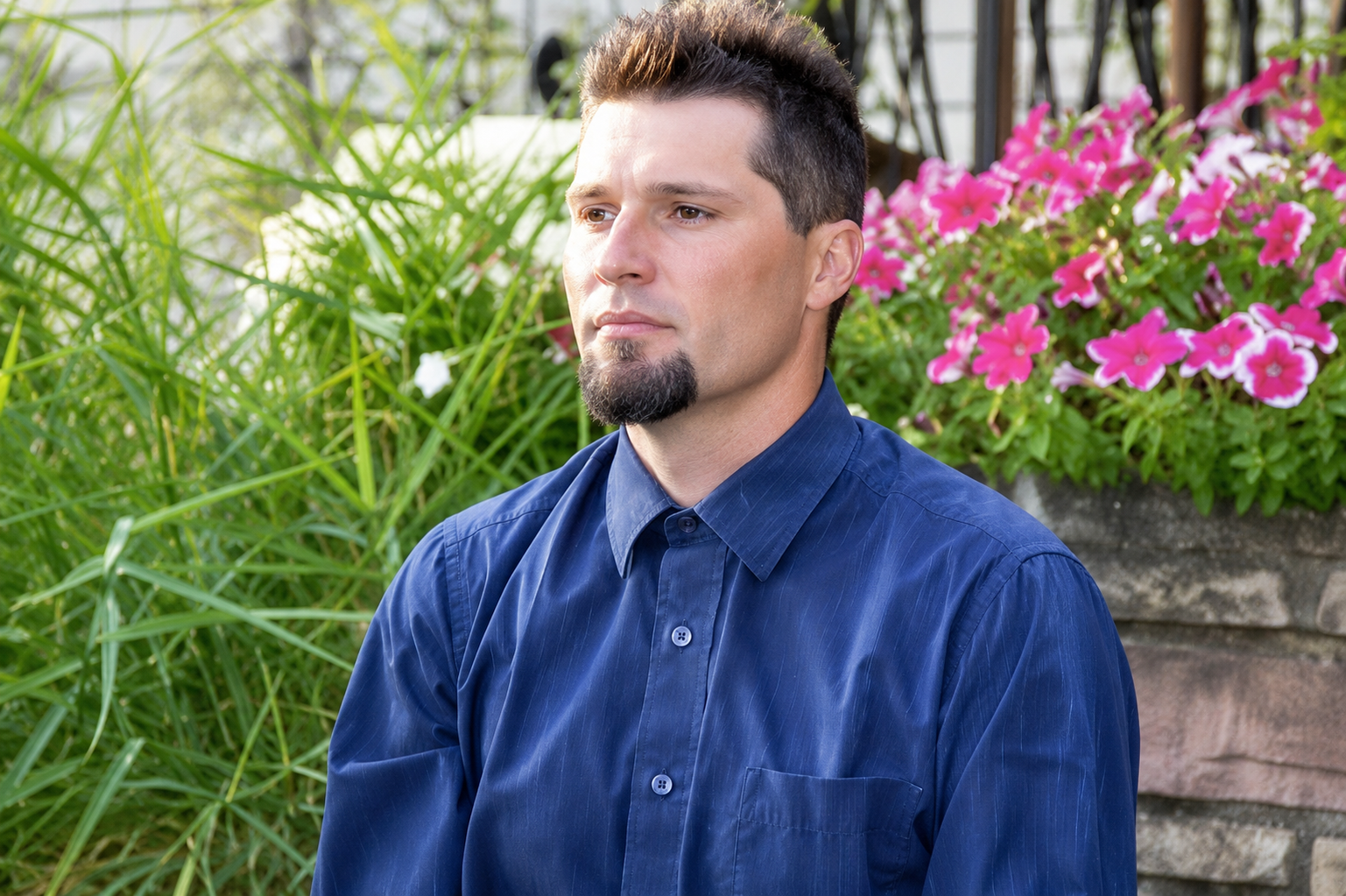 Man in a blue shirt standing outdoors beside pink flowers and greenery in Columbus Ohio