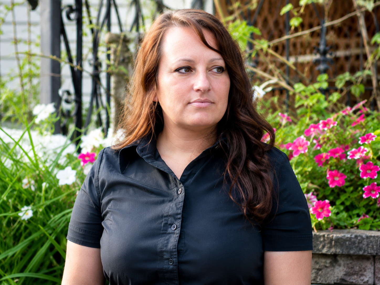 Woman standing outdoors near pink flowers and a fence, wearing a dark shirt, looking to the side in Columbus Ohio