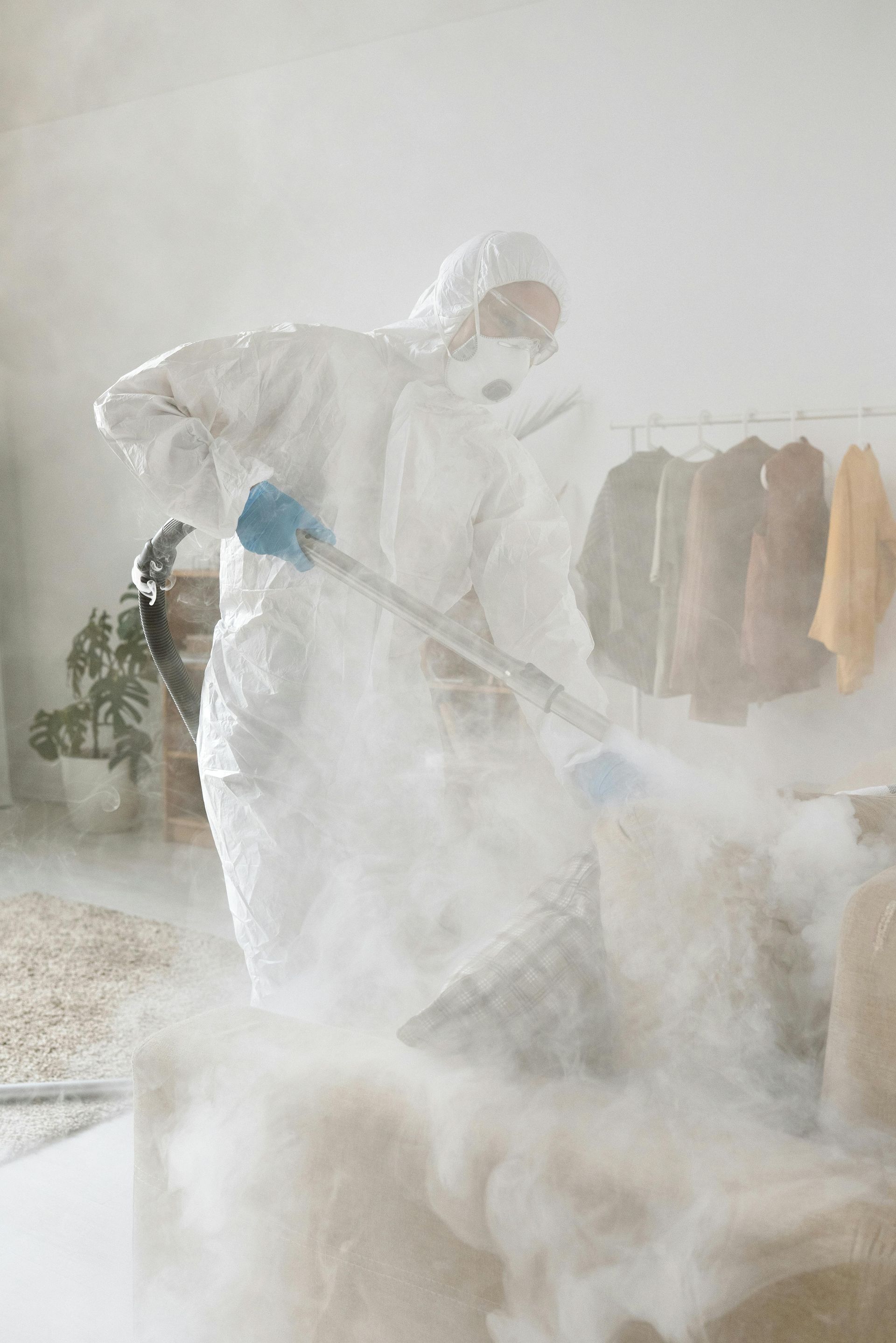 Person in white protective suit cleaning with a spray in a hazy room, with hanging clothes in the background in Parkersburg