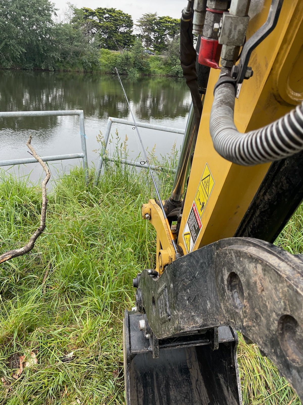 Yellow Excavator Near Water And Grass.