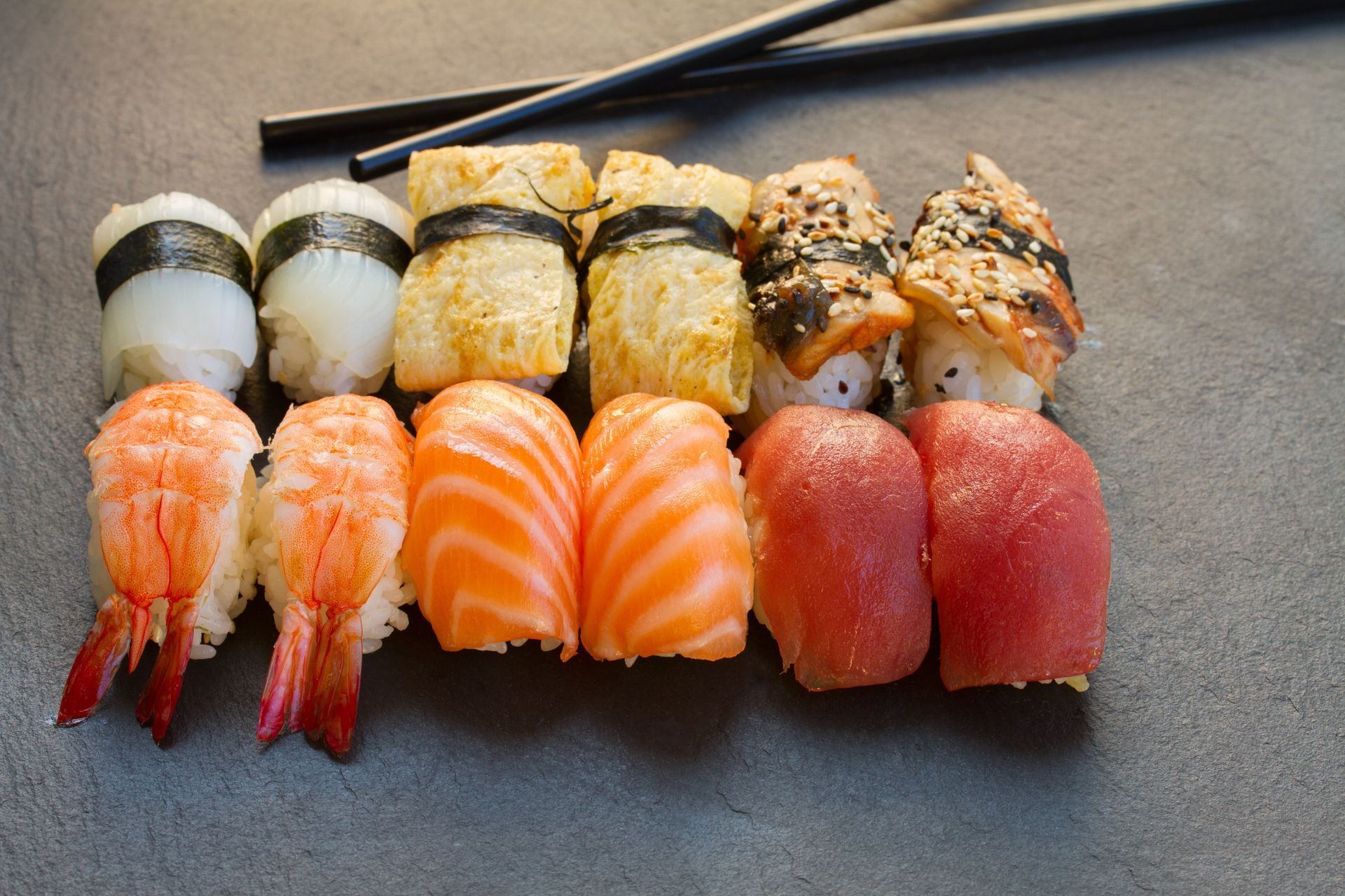Assorted sushi nigiri and maki arranged on a dark tray with chopsticks above.