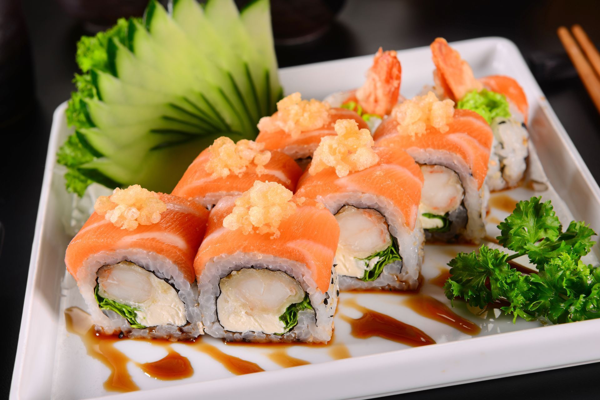 Sushi rolls topped with salmon on a white plate, garnished with greens and sauce